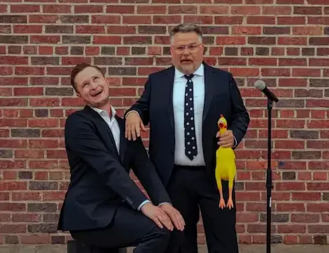 Donovan and Chris sitting in front of a brick wall with a rubber chicken.